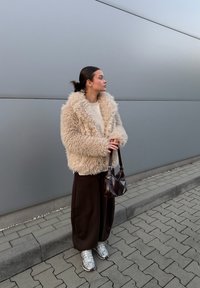 Beige shaggy faux fur jacket over a cream jumper, paired with brown wide-legged trousers and silver trainers. Dark brown shoulder bag.