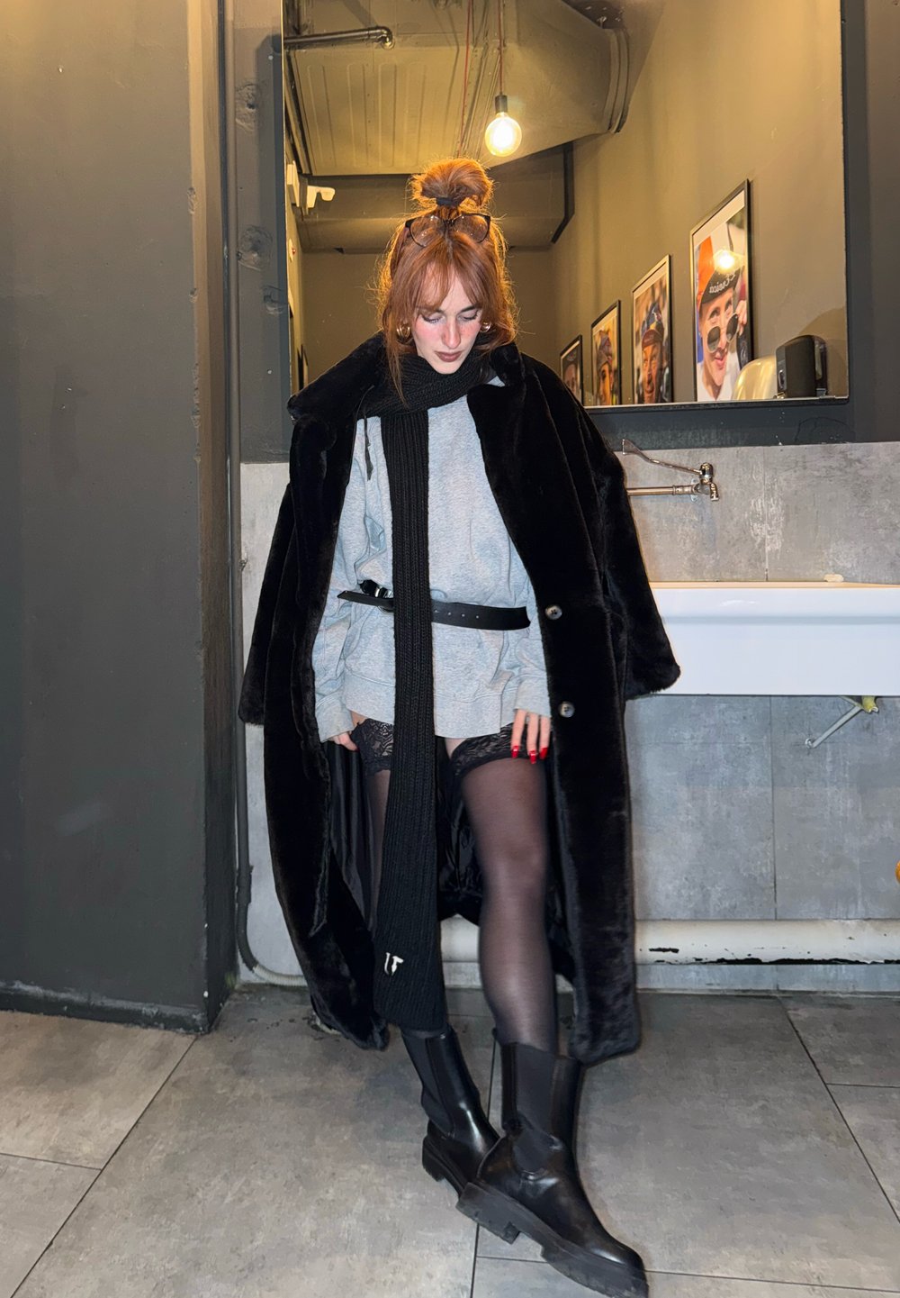 Čierny kabát z umelej kožušiny, sivý oversize sveter, čierne pančuchy s čipkovým detailom a čierne členkové topánky. Dlhá čierna šál, červené nechty. Pozadie s osvetlením.