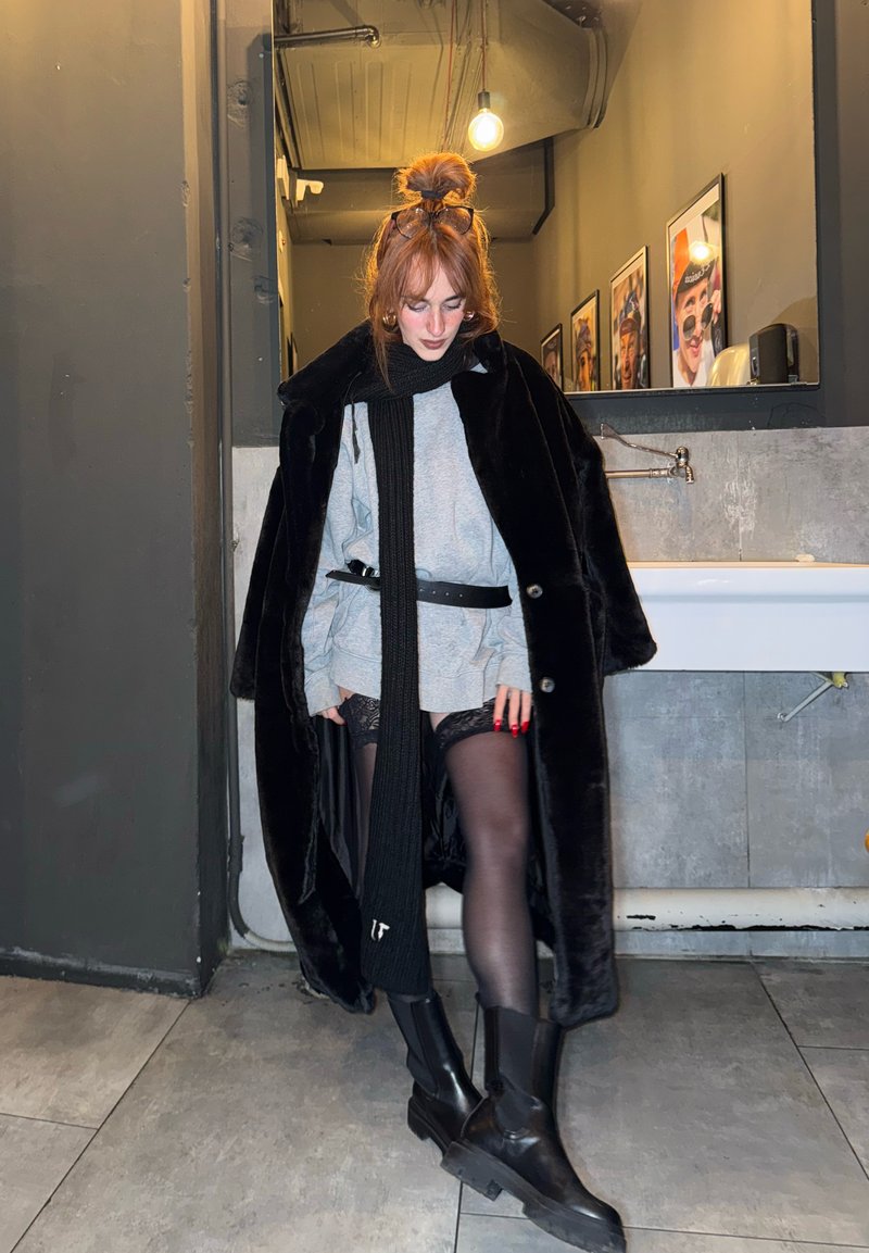 Black faux fur coat, gray oversized sweater, black tights with lace detail, and black ankle boots. Long black scarf, red nails. Background with light.