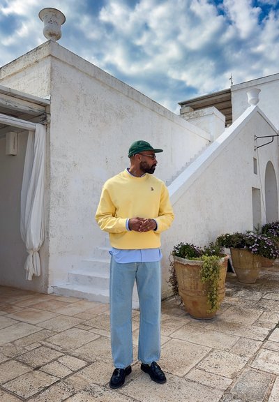 Hombre con sudadera amarilla, camisa azul, vaqueros claros y gorra verde, de pie en un patio de piedra cerca de un edificio blanco y grandes plantas en macetas.