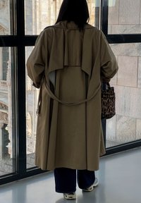 Beige trenchcoat med draperad bakdel, i kombination med mörka vida jeans och en brun leopardmönstrad väska. Inomhusmiljö med stora fönster.