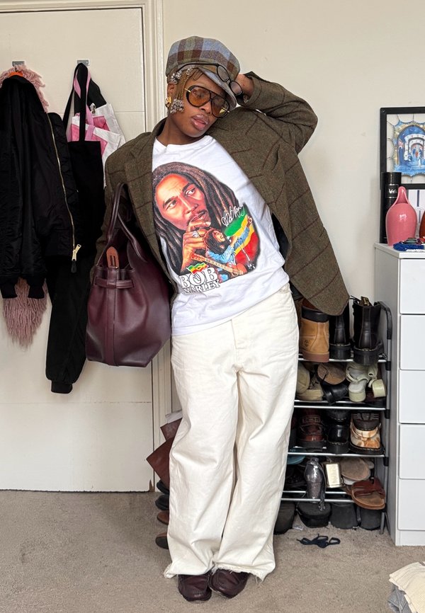 Personne portant un blazer à carreaux marron, un t-shirt graphique Bob Marley, un pantalon blanc, de grandes lunettes et un chapeau, appuyée avec un sac marron dans une pièce avec des chaussures.