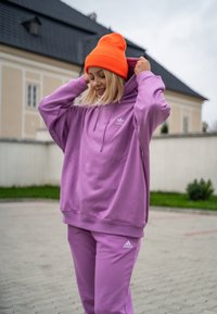 Violet huppari ja yhteensopivat verkkareita pehmeästä kankaasta, yhdistettynä oranssiin neulottuun päänmyssyyn. Adidas-logo hupparin ja housujen edessä.