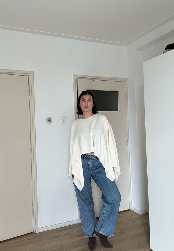 Femme debout à l'intérieur portant un haut asymétrique blanc ample, un jean bleu à jambes larges et des bottines foncées devant un mur uni et des portes fermées.