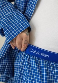 Chemise à carreaux bleue Calvin Klein avec ceinture, présentant un motif blanc et un logo, avec un gros plan sur une main affichant des tatouages.