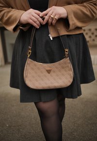 Femme portant une veste marron et une jupe foncée, tenant un sac à main beige Guess avec un motif de logo sur la bandoulière.