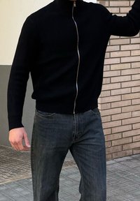 Personne portant un pull noir côtelé à fermeture éclair et un jean foncé, debout sur un trottoir carrelé devant un mur en briques beige.