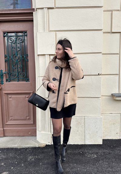Chaqueta de forro polar beige con acentos negros y cierres de toggles, llevada sobre un vestido negro, combinada con botas negras hasta la rodilla y un bolso negro.
