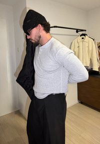 Chemise grise à manches longues, pantalon noir et veste noire tenue sur l'épaule. Accessoires comprennent un bonnet noir et des lunettes de soleil. Sol en bois visible.