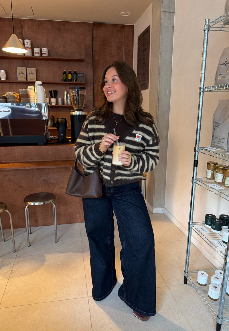 Cardigan a righe marrone scuro e crema, jeans a gamba larga scuri, tiene in mano una bevanda ghiacciata, in piedi in un caffè con elementi in legno e metallo.