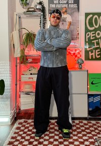 Homme portant un pull à col roulé gris, un pantalon noir, un bandana et des lunettes de soleil, debout les bras croisés dans une pièce avec des étagères à baskets et des affiches.