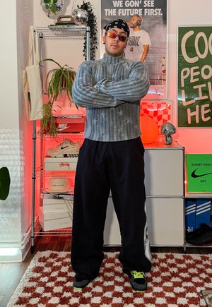 Hombre con suéter de cuello alto gris, pantalones negros, bandana y gafas de sol, de pie con los brazos cruzados en una habitación con estantes de zapatillas y carteles.