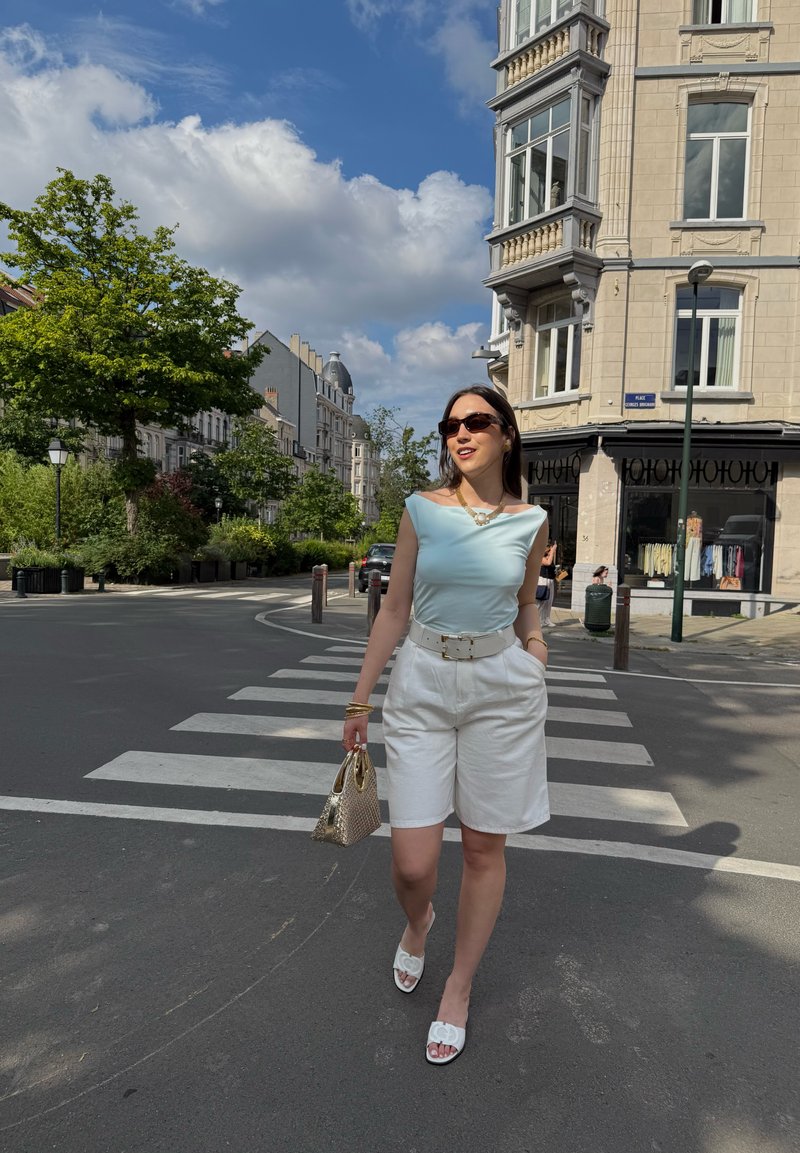 Femme portant des lunettes de soleil et un short blanc traverse un passage piéton en ville par une journée ensoleillée, avec des arbres et des bâtiments historiques en arrière-plan.