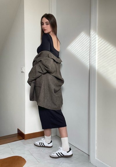 Abrigo de lana marrón oversize sobre una blusa negra ajustada, combinado con una falda midi negra y zapatillas Adidas blancas con rayas negras.