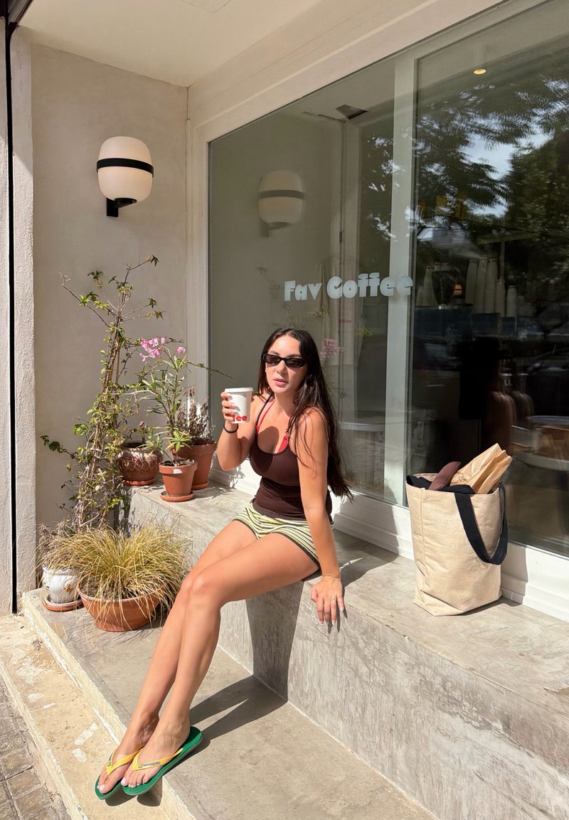 Femme avec des lunettes de soleil tenant une tasse de café assise à l'extérieur d'un café à côté de plantes en pot et d'un sac contenant des baguettes.