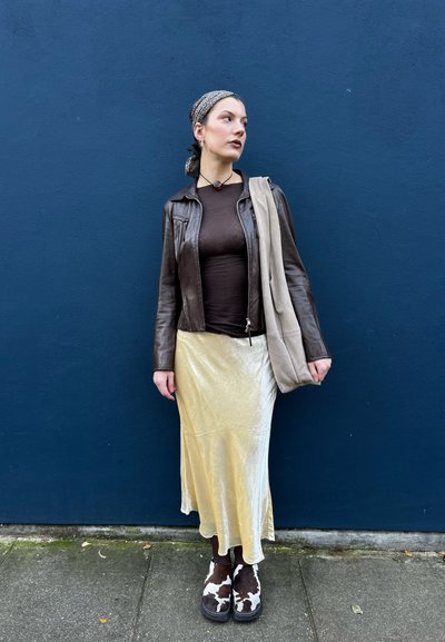 Chaqueta de cuero marrón, blusa marrón ajustada, falda larga de satén dorado, zapatos de plataforma con un patrón marrón y blanco, llevando un bolso de hombro beige.