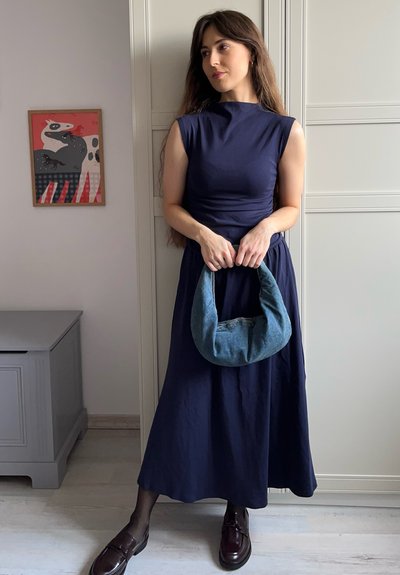 Un vestido sin mangas de color azul marino con un cinturón fruncido, combinado con un bolso de cuero vaquero texturizado. Unos zapatos oscuros y pulidos completan el conjunto.