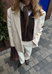 Manteau blanc texturé porté sur un pull marron, associé à un pantalon blanc et des chaussures marron foncé. Une écharpe à motifs et un sac marron complètent le look.