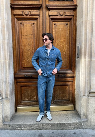 Chaqueta de denim con dos bolsillos frontales, combinada con jeans a juego y zapatillas blancas. Las gafas de sol completan el look. Fondo de puerta de madera.