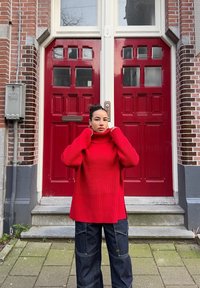 Maglione a collo alto a coste rosso con un design lungo e morbido abbinato a pantaloni a gamba larga in denim scuro, in piedi davanti a porte doppie rosse.