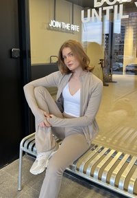 Tenue de sport beige composée d'une veste ajustée à fermeture éclair et d'un legging assorti, portée avec des baskets blanches, assise sur un banc argenté.
