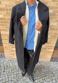 Persona che indossa una camicia blu con bottoni d'argento, pantaloni di pelle nera, scarpe nere a punta e un lungo cappotto nero tenuto aperto su una strada di ciottoli.