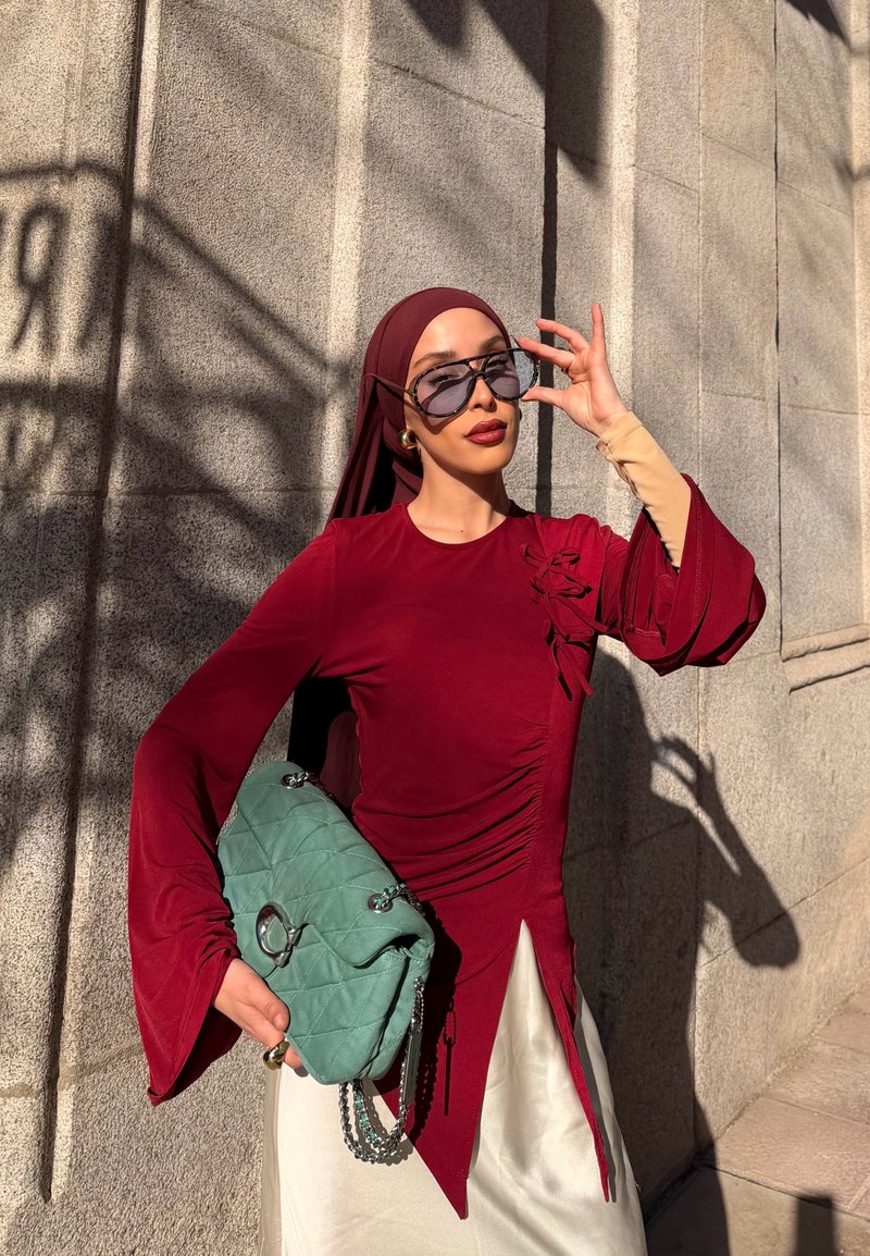 Vrouw in wijnrode jurk en hoofddoek die haar zonnebril rechtzet en een groene doorgestikte handtas vasthoudt, staand tegen een zonovergoten stenen muur.