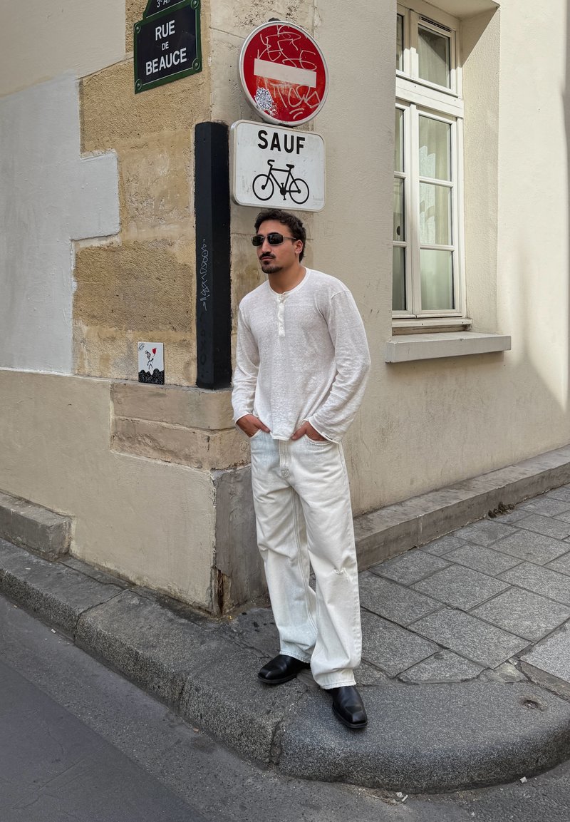 Homme portant une chemise blanche et un pantalon avec des chaussures noires et des lunettes de soleil se tient au coin de la rue près des panneaux d'interdiction d'entrée et de vélo.