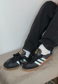 Zwarte suede sneakers met blauwe strepen, voorzien van een gumrubberen zool en een gele logotag, gecombineerd met donkere spijkerbroeken en witte sokken.