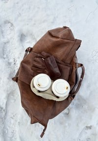 Sac en cuir marron sur la neige avec des gants en cuir, un portefeuille et deux gobelets de café à emporter dans un porte-gobelets en carton posés dessus.