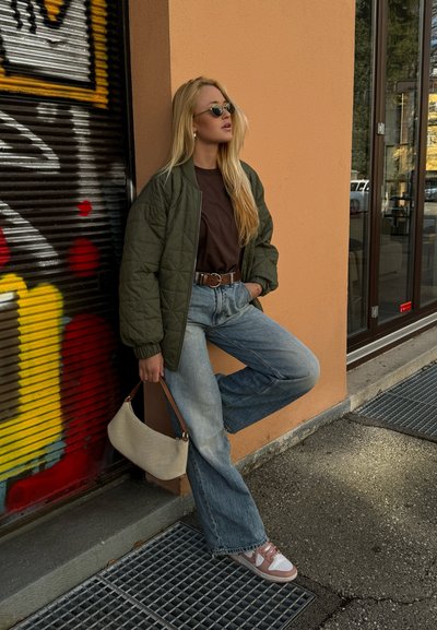 Chaqueta bomber acolchada verde oliva sobre una blusa marrón, combinada con pantalones de mezclilla de pierna ancha de color azul claro y zapatillas rosa y blancas, sosteniendo un bolso beige.