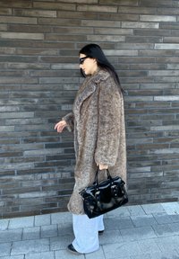 Μακρύ καφέ γούνινο παλτό από συνθετική γούνα με υπερμεγέθη μανίκια, συνδυασμένο με γκρίζες παντελόνες και μαύρη δερμάτινη τσάντα χειρός με ασημένιες λεπτομέρειες.
