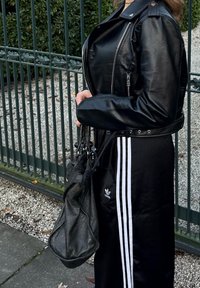 Veste courte en cuir noir avec des fermetures éclair argentées, associée à un pantalon noir avec des bandes blanches et un logo Adidas. Sac noir à la main.