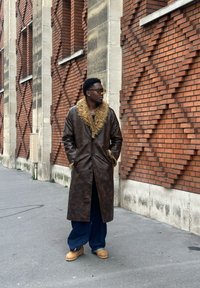 Casaco longo de couro castanho com colarinho de pelo, combinado com calças azuis largas e botas castanhas claras. Em pé contra uma parede de tijolos com um padrão texturizado.