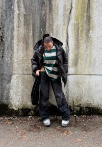 Doudoune noire, t-shirt rayé vert et blanc, jean noir ample, baskets argentées, portant un sac noir, devant un mur texturé.