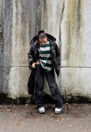 Chaqueta acolchada negra, camiseta a rayas verdes y blancas, vaqueros negros holgados, zapatillas plateadas, lleva un bolso negro, contra una pared texturizada.