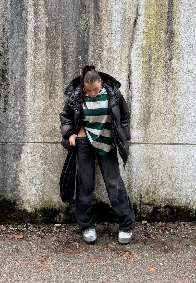 Veste matelassée noire, t-shirt rayé vert et blanc, jean noir ample, baskets argentées, portant un sac noir, devant un mur texturé.