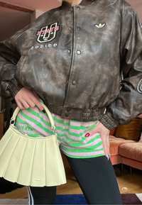 Personne portant une veste bomber Adidas marron, un short à rayures rose et vert, des leggings noirs, tenant un sac à main plissé crème à l'intérieur.