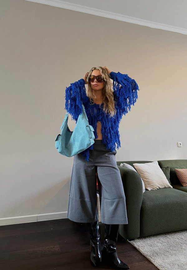 Femeie purtând pulover albastru cu franjuri, pantaloni gri cu croială largă, cizme negre, ținând o geantă albastru deschis, stând în camera de zi cu o canapea verde și perne.