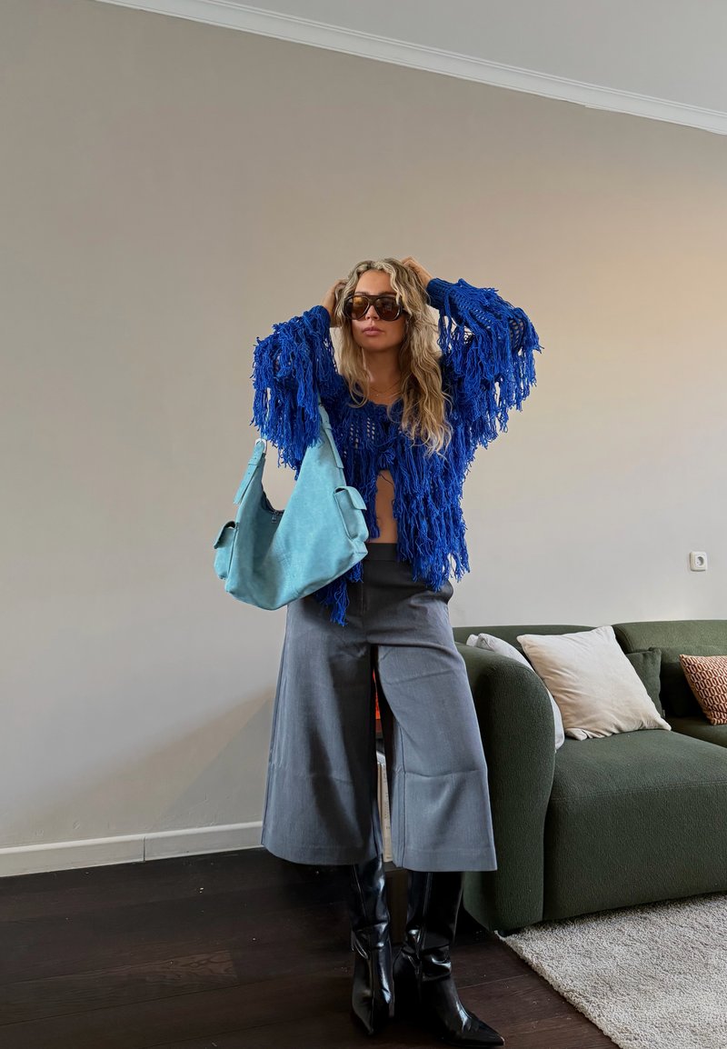 Γυναίκα που φοράει μπλε πουλόβερ με φούντες, γκρι φαρδιά παντελόνια, μαύρες μπότες, κρατάει γαλάζια τσάντα, στέκεται στο σαλόνι με πράσινο καναπέ και μαξιλάρια.