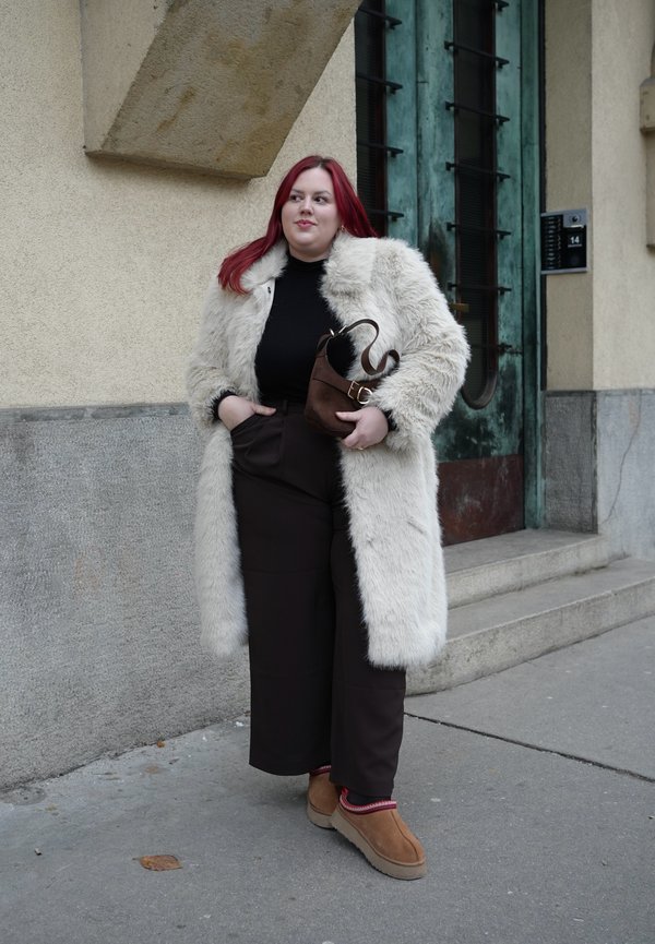 Punase juuksevärviga inimene, kes kannab valget karvkattega mantlit, musta topi, tumedaid laiade säärega pükse ja pruunikaid kingi, hoides pruunist käekotti hoone sissekäigu juures.