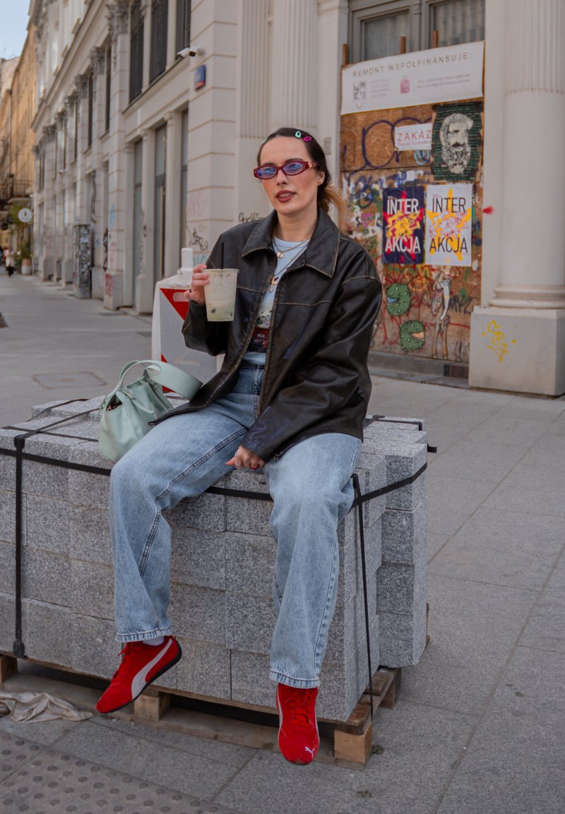 Žena v černé kožené bundě a červených teniskách sedí na kamenných blocích, drží nápoj, za ní je zeď pokrytá graffiti.