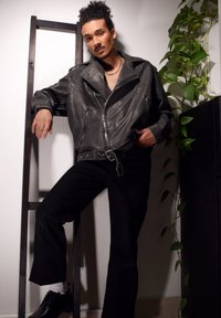 Veste de moto en cuir noir avec des zippers argentés, associée à un pantalon large noir et des chaussures noires. Plante verte en arrière-plan.