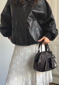 Casaco oversized em pele preta com gola com fecho de correr, combinado com uma saia branca texturada em renda e uma mala de mão em pele castanha.