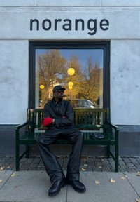 Hombre con traje de cuero negro y guantes rojos sentado en un banco verde fuera de un edificio con un letrero que dice "norange" y una ventana que refleja árboles.