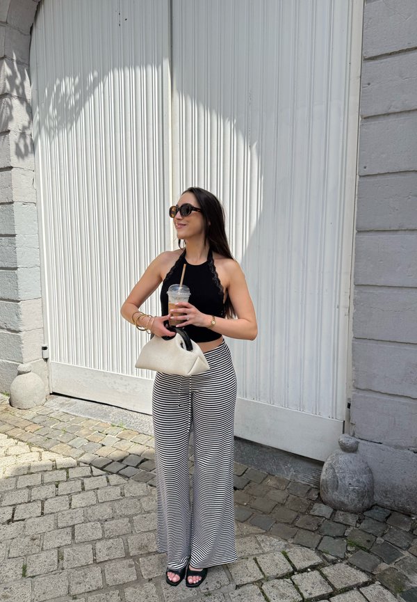 Žena s okuliarmi na slnko, čiernym tielkom bez rukávov, pruhovanými širokými nohavicami, drží bielu kabelku a plastový pohár so slamkou vonku.