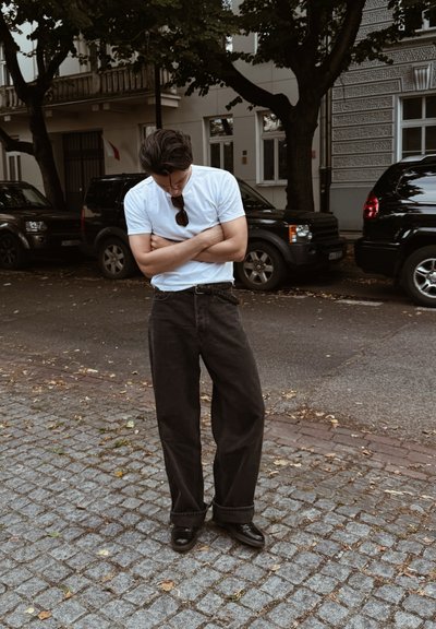 Hombre con camiseta blanca y pantalones anchos negros se encuentra de pie en una calle de adoquines con los brazos cruzados y la cabeza inclinada, cerca de coches aparcados y árboles.
