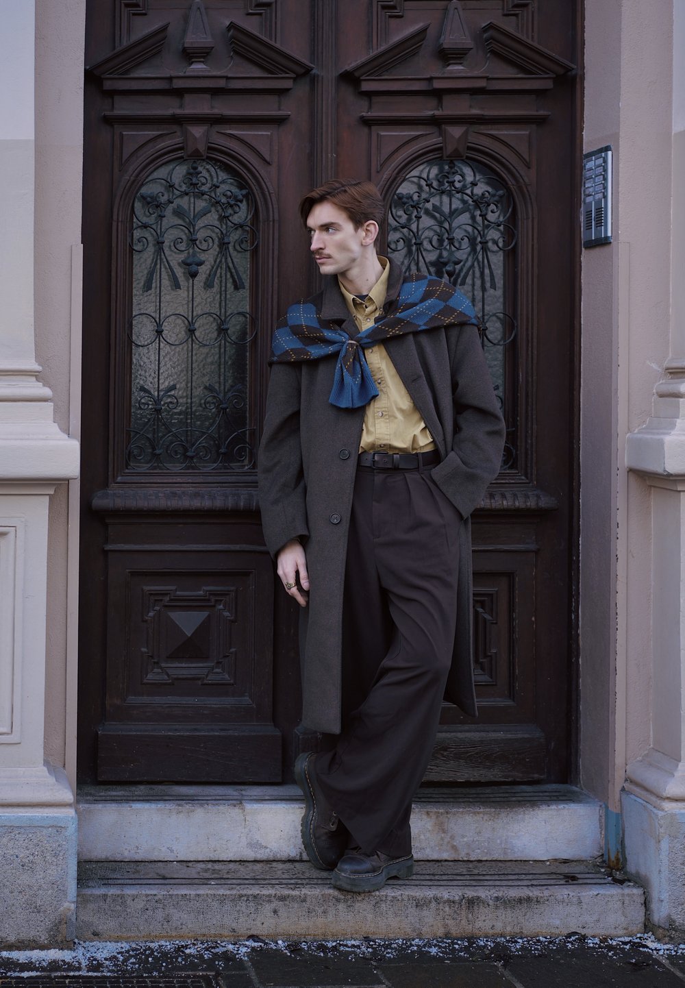 Jeune homme à moustache portant un long manteau foncé, une chemise beige, un pantalon sombre et une écharpe bleue à motifs, appuyé contre une porte en bois ornée à l'extérieur.