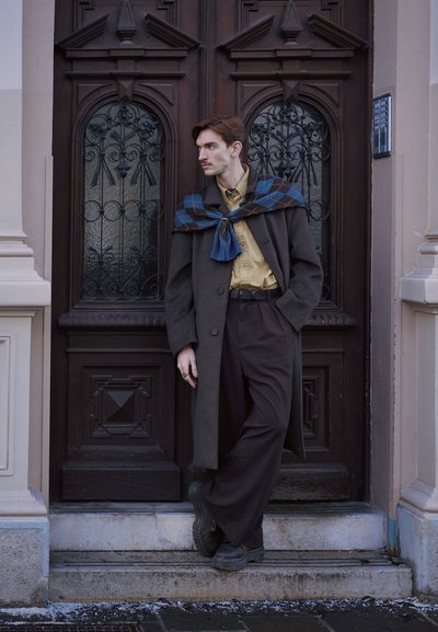 Joven con bigote que lleva un abrigo largo oscuro, camisa beige, pantalones oscuros y una bufanda azul con estampado, apoyado en una puerta de madera ornamentada al aire libre.