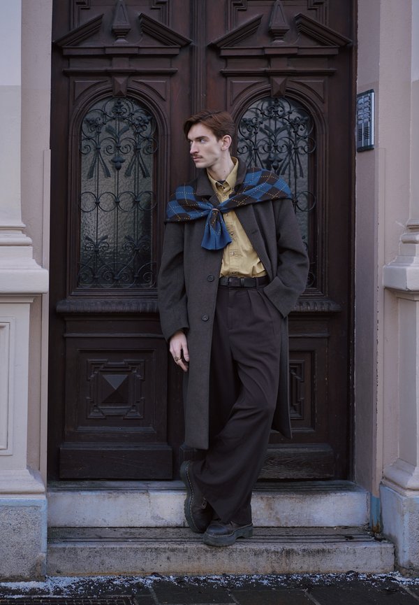 Ung mann med bart iført lang mørk frakk, beige skjorte, mørke bukser og blå skjerf med mønster, lent mot en utsmykket tredør utendørs.